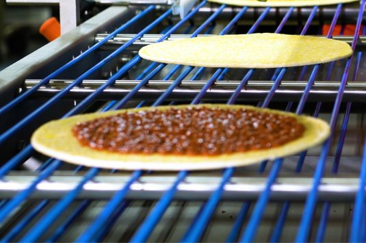 A pizza in production having sauce applied from a sauce applicator. 